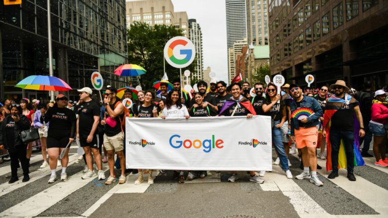 Google marching in the Toronto Pride parade in 2024. A crowd holds rainbow umbrellas and fans, a Google banner and a placard with a Google logo