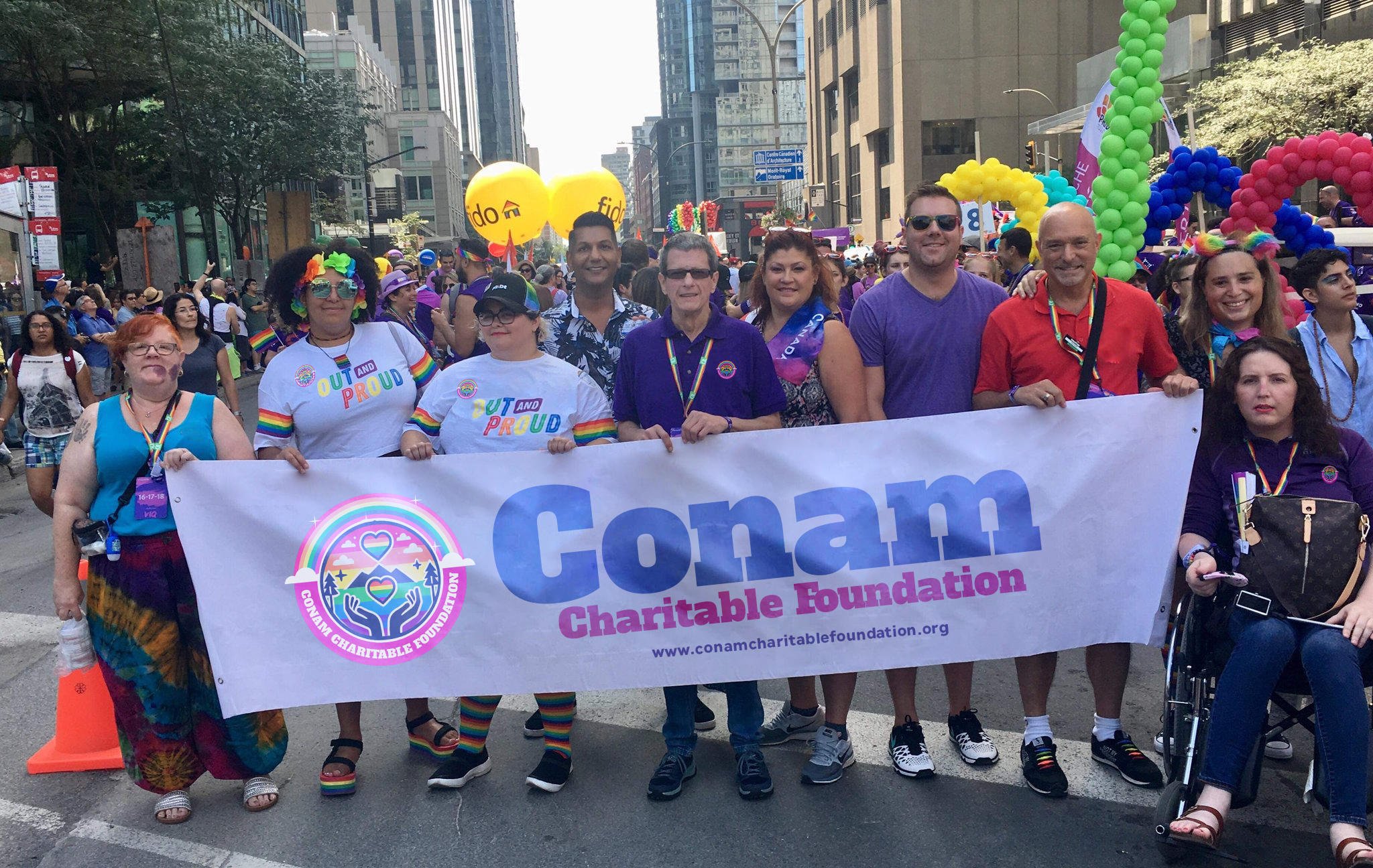 People marching in a Pride Parade with a banner that says "Conam Charitable Foundation" with a URL underneath