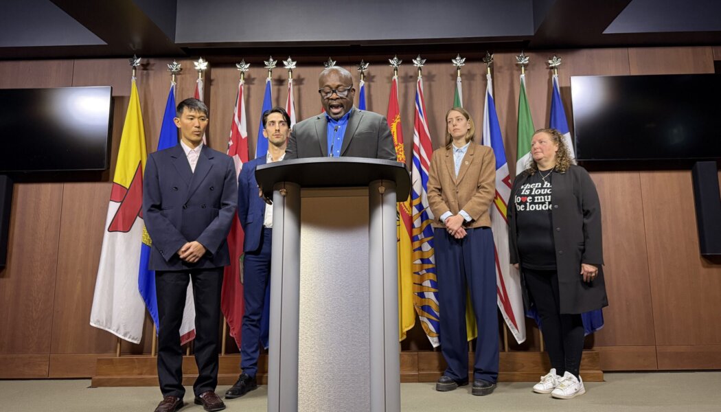 Five leaders of Canadian Pride organizations stand behind a podium with a row of flags behind them