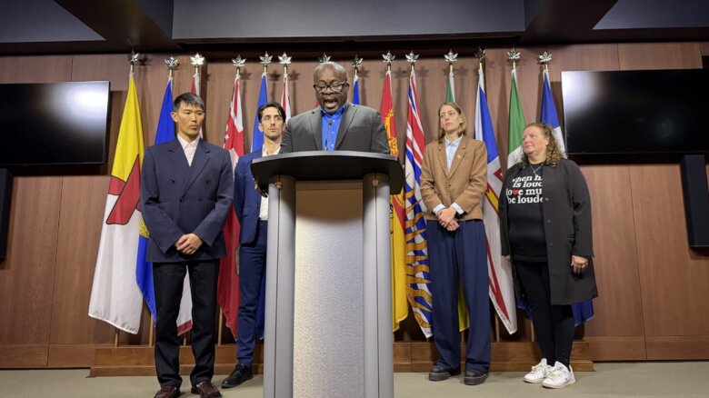 Five leaders of Canadian Pride organizations stand behind a podium with a row of flags behind them