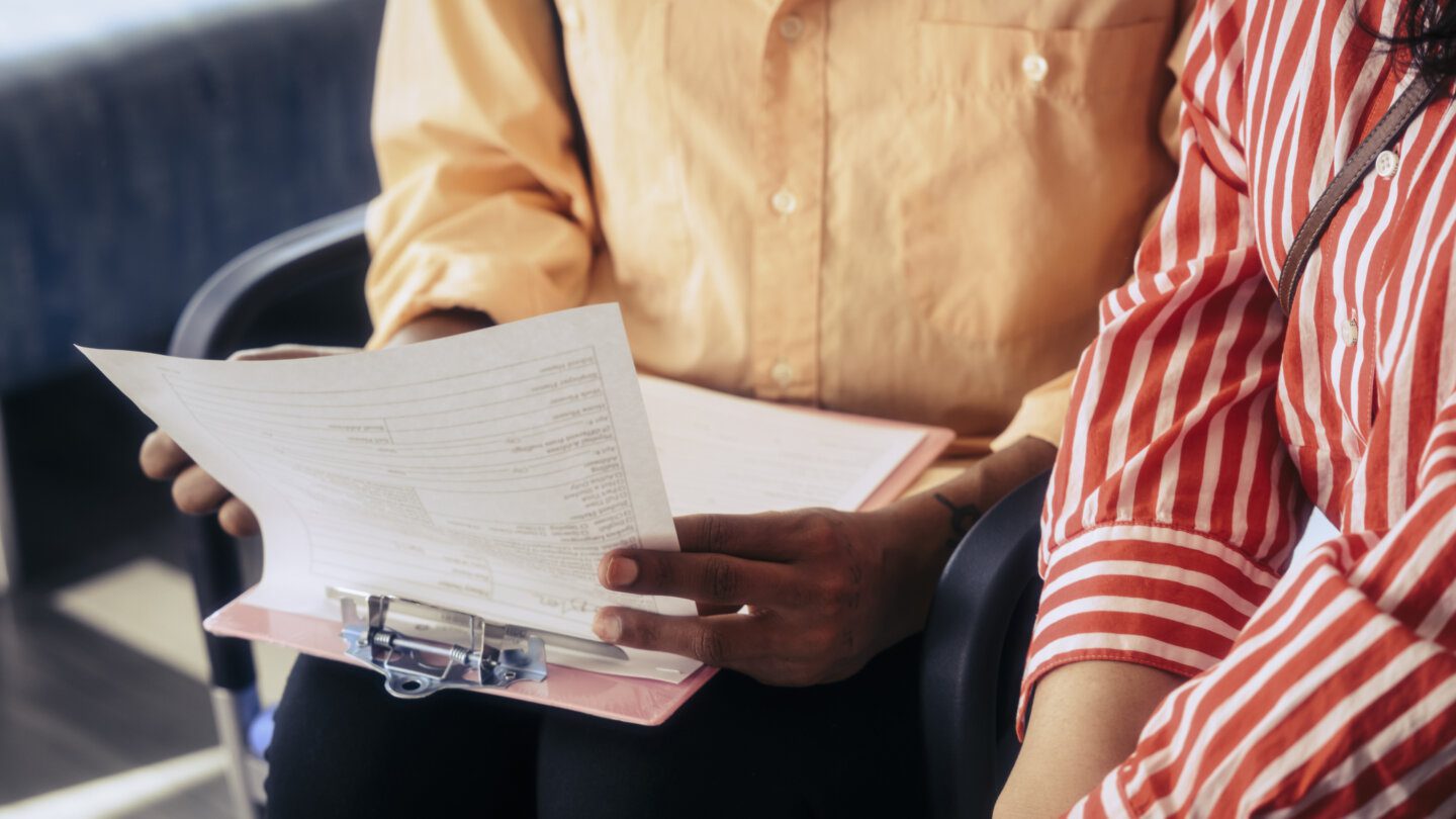 Midsection of person holding clipboard with forms, sitting next to another person
