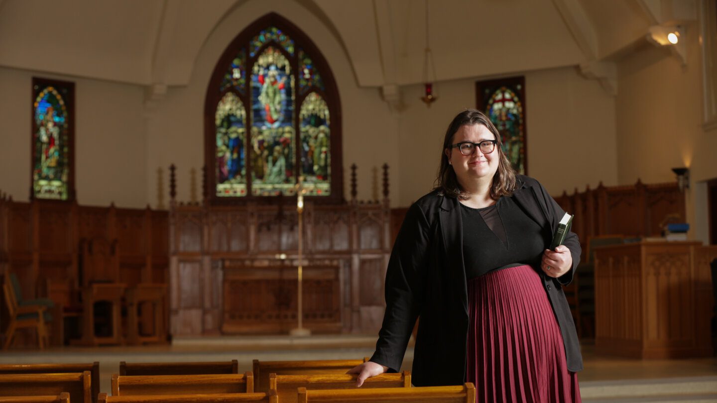 Author Charlotte Sheasby wears a black top and blazer and maroon pleated skirt. She has dark-framed glasses and shoulder-length brown hair. She holds a bible, posing inside of St. Stephen's Anglican.