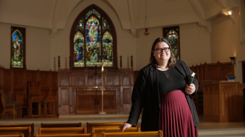 Author Charlotte Sheasby wears a black top and blazer and maroon pleated skirt. She has dark-framed glasses and shoulder-length brown hair. She holds a bible, posing inside of St. Stephen's Anglican.