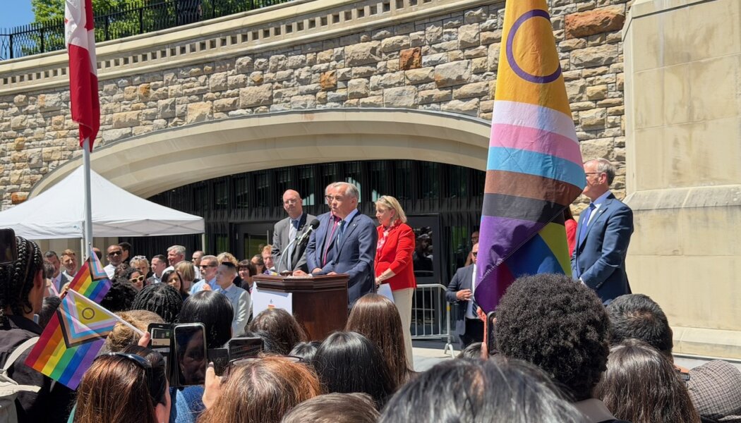 Mark Carney stands at a podium between a Progress Pride flag and Canadian flag. A seated crowd faces him.
