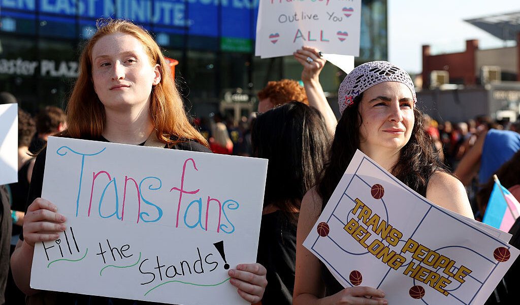 Two counter-protesters holding signs that say "Trans fans fill the stands!" and "Trans people belong here." A trans flag and a sign that says "Trans joy will outlive you all" are also visible.