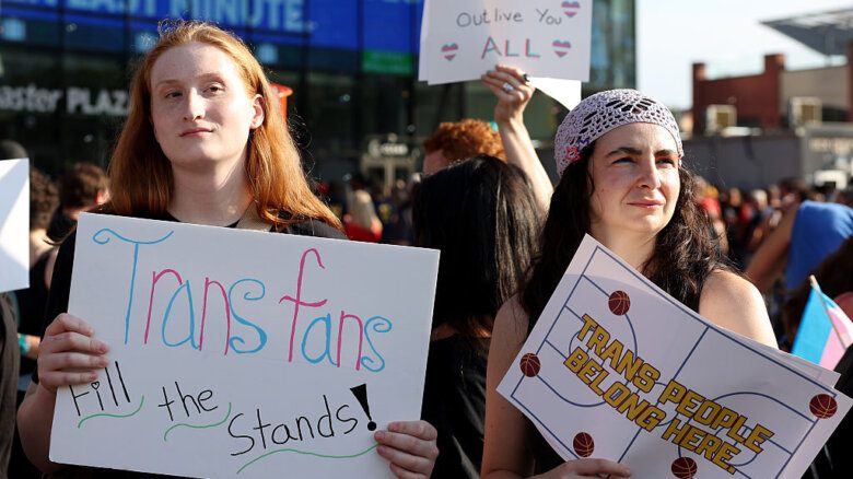 Two counter-protesters holding signs that say "Trans fans fill the stands!" and "Trans people belong here." A trans flag and a sign that says "Trans joy will outlive you all" are also visible.