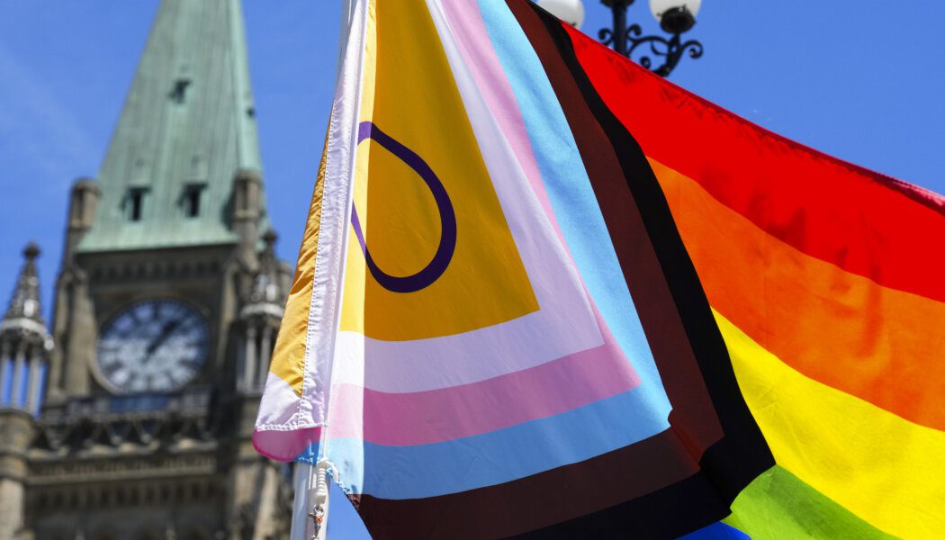 A Pride flag is shown close up in front of Parliament Hill, which is in the background