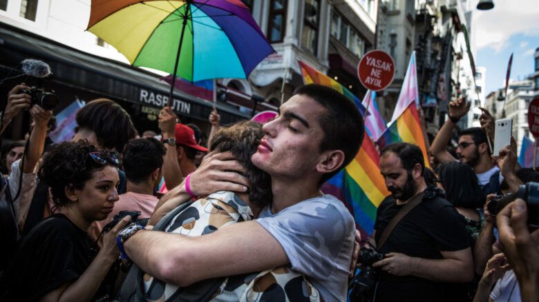 Two people embrace amongst Pride flags and rainbow umbrellas.