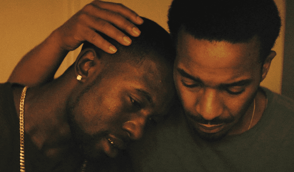 Two Black men are shown in the image; one has his head on the other's shoulder; the other appears to gently cusp the other's head in his hand